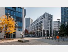 Städtischer Platz mit modernen Bürogebäuden aus hellem Stein und vielen Glasfenstern; im Vordergrund stehen herbstlich gefärbte Bäume, eine Frau mit Kamera, ein moderner Brunnen und ein orangefarbener Briefkasten. Die Straße im Hintergrund ist von weiteren Gebäuden und parkenden Autos gesäumt.