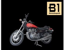 Ein rotes, klassisches Motorrad der Marke Kawasaki mit einem schwarzen Sitz und verchromten Details vor schwarzem Hintergrund. Rechts oben ist ein Schild mit der Aufschrift „B1 Gewerbepark“ auf beigem Hintergrund sichtbar.