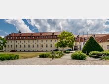 Historisches Gebäude mit hellgelber Fassade und rotem Satteldach, flankiert von gepflegten Grünflächen mit Beeten, einem Baum in der Mitte und einer Hecke in Form eines Kegels; vorne führt ein gepflasterter Weg vorbei an einer Bank. Der Himmel ist von Wolken durchzogen.