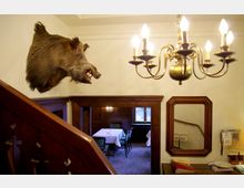 Treppenhaus eines rustikalen Gasthauses mit Holzgeländer, Wanddekorationen wie einem ausgestopften Wildschweinkopf und einem großen, goldenen Kronleuchter. Im Hintergrund ein Raum mit gedeckten Tischen und gepolsterten Stühlen.