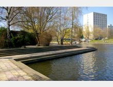 Ein Uferbereich mit breiten Stufen und einem gepflasterten Weg, der zu einem ruhigen Gewässer führt, von Bäumen und Sträuchern umgeben. Im Hintergrund steht ein mehrstöckiges Gebäude neben einer Fußgängerbrücke, die das Wasser überquert.