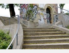 Außentreppe aus Beton mit Metallgeländern, die zu einem graffiti-bedeckten Gebäude mit einer Bogenöffnung führt; umgeben von Bäumen und Pflanzen.