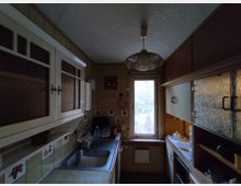 Schmale, altmodische Küche mit braunen und cremefarbenen Fliesenwänden, einer Spüle auf der linken Seite und Hängeschränken mit verziertem Glas. In der Mitte befindet sich ein kleines Fenster mit Blick ins Freie, darunter ein Heizkörper; rechts sind weitere Schränke und ein kleiner Herd eingebaut.