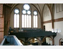 Innenraum einer historischen Kirche mit hohen, dekorativen Buntglasfenstern und gemauerten Rundbögen. Dunkle Holzempore mit Sitzbänken und modernen Hängelampen, umgeben von Backstein- und Putzwänden mit verzierten Elementen.