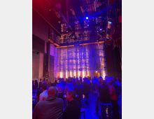 Eine Veranstaltungsbühne in einem modern gestalteten Saal mit hohen Wänden, die durch warme Beleuchtung und sichtbare Metallstrukturen betont werden. Im Vordergrund sind mehrere Personen auf Stühlen und Stehtischen in einem abgedunkelten Raum zu sehen.