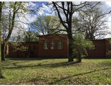 Backsteingebäude mit großen Bogenfenstern in einer parkähnlichen Umgebung mit grüner Wiese, hohen Bäumen und vereinzelten Sträuchern im Vordergrund. Der Himmel ist teils bewölkt mit Sonnenlicht, das durch die Bäume fällt.