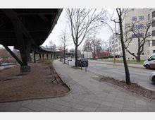 Straßenszene in einer städtischen Umgebung mit einem Fußweg, Autos auf der Straße und modernen Gebäuden im Hintergrund. Links im Bild ist eine erhöhte, stählerne Bahntrasse zu sehen, die parallel zu einem Kanal verläuft.