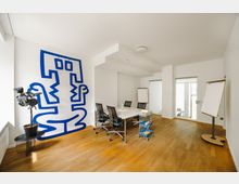 Heller Besprechungsraum mit Holzboden, einem rechteckigen Tisch mit Bürostühlen und einem großen blauen Wandbild. An den Wänden befinden sich zwei Flipcharts, und ein kleiner Rollwagen mit Schreibmaterialien steht neben dem Tisch.