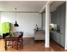 Offener Wohnraum mit einer modernen Küche in grauen Tönen auf der rechten Seite und einem Essbereich mit Holztisch und bunten Stühlen auf der linken Seite. Der Raum hat eine weiße Wand, einen hölzernen Boden sowie eine auffällige grüne Wanddekoration und eine schwarze Hängelampe über dem Tisch.