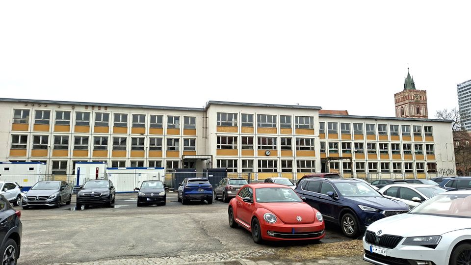 Großer, rechteckiger Schul- oder Verwaltungsbau mit drei Stockwerken, regelmäßiger Fensterfront und beigefarbenen Fassadenfeldern. Davor liegt ein asphaltierter Parkplatz mit mehreren Autos sowie Baustellencontainern und Absperrzäunen; im Hintergrund ragt ein Kirchturm über das Gebäude.
