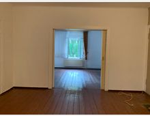 Ein leerer Raum mit Holzboden und weißen Wänden, der durch eine breite, offene Tür in einen weiteren Raum führt. Im hinteren Raum befindet sich ein großes Fenster mit weißen Vorhängen, durch das Bäume sichtbar sind, sowie ein Heizkörper unter dem Fenster.