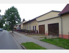 Straßenszene in einem Wohngebiet mit gelb verputzten Reihenhäusern, kleinen Vorgärten und aneinander angrenzenden Holztoren. Links verläuft eine asphaltierte Straße mit Bäumen und Gehweg, am Horizont sind weitere Häuser und Fahrzeuge zu sehen.