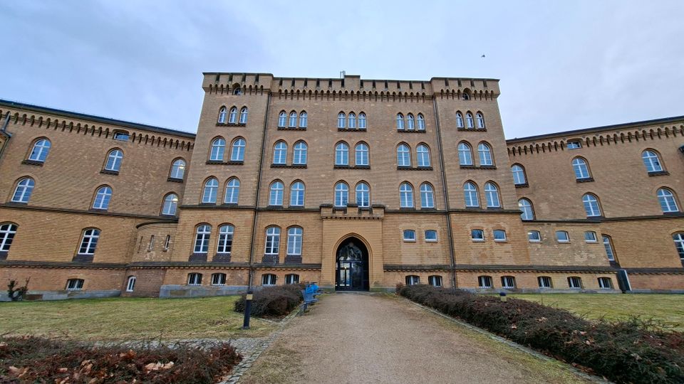 Frontansicht eines großen, mehrstöckigen Backsteingebäudes mit vielen symmetrisch angeordneten Rundbogenfenstern und einem mittigen Spitzbogen-Eingang. Ein gerader Kiesweg führt durch eine Rasenfläche mit niedrigen Hecken zur Eingangstür unter bewölktem Himmel.