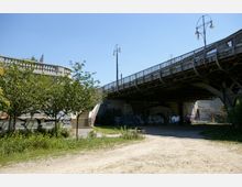 Stadtlandschaft unter einer Brücke mit Stahlträgern und Laternen, umgeben von Bäumen und Grünflächen. Graffiti bedecken die Betonflächen der Brückenstützen, und an der linken Seite führt eine Treppe nach oben.