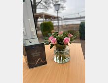 Ein heller Holztisch mit einer Glasvase voller rosa Blumen und Grünzeug steht im Vordergrund. Daneben liegt eine aufrechte Getränkekarte, im Hintergrund sind unscharf ein Außensitzbereich, Bäume und ein Gewässer zu erkennen.