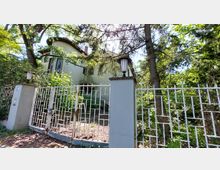 Ein wohnhausartiges Gebäude mit einem gewölbten Dach steht hinter einer weißen Metallzaun-Konstruktion und ist von hohen Bäumen und viel Grün umgeben. Zwei Zaunpfeiler mit Laternen flankieren den Eingang, der über einen kleinen Weg zugänglich ist.