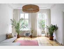 Wohnzimmer mit heller Einrichtung, großen Fenstern und Zugang zu einem Balkon. Der Raum ist mit Pflanzen, einem hellgrauen Sofa, einem rosafarbenen Teppich und einem auffälligen Lampenschirm aus Naturmaterialien dekoriert.
