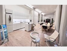 Helles, modern eingerichtetes Wohnzimmer und offene Küche mit weißen Einbauschränken, Holzfußboden und langen weißen Vorhängen. Im Raum stehen ein Esstisch mit Stühlen, ein kleiner Tresen mit Barhockern und verschiedene Sitzmöbel, dekoriert mit schlichten Accessoires.