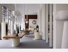 Innenansicht eines modernen Showrooms mit skulpturalen Möbeln und Dekorationen. Der Raum ist durch hohe Fenster, weiße Wände mit Säulen und elegante Holztische mit geschwungenen Formen geprägt; mannequinförmige Ständer und Objekte setzen Akzente.