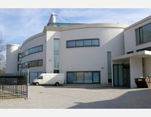 Modernes Gebäude mit geschwungener Fassade, weißen Wänden und großen, schmalen Fenstern. Im Vordergrund ein gepflasterter Hof mit einem weißen Lieferwagen und einem metallenen Zaunrand.