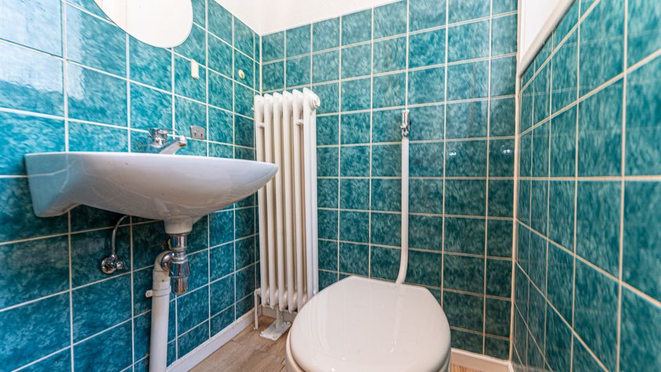 Kleines Badezimmer mit türkisfarbenen Wandfliesen, wandmontiertem Waschbecken und WC. An der Rückwand steht ein weißer Heizkörper; der Boden ist in Holzoptik gehalten.