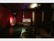 Small indoor music venue with a low stage in front of red curtains, featuring a white upright piano and an acoustic guitar near stage lighting. Wooden floors, scattered chairs and tables, and ceiling-mounted spotlights with colored light reflections define the intimate performance space.