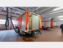 Eine Fahrzeughalle mit mehreren geparkten Feuerwehrfahrzeugen in orange-weißer Lackierung. Sichtbar sind Abgasschläuche, die an den Fahrzeugen angeschlossen sind, sowie eine helle, offene Decke mit sichtbaren Rohrleitungen und Beleuchtung.