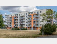 Mehrstöckiges Wohngebäude mit hellbeiger Fassade, orangen Akzenten und zahlreichen Balkonen. Im Vordergrund befindet sich eine Wiese mit vereinzelten Bäumen und Sträuchern, sowie ein Gehweg mit einer Straßenlaterne.