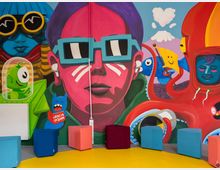 Innenraum mit einer großflächigen, bunten Wandmalerei im Pop-Art-Stil, die stilisierte Figuren und Comicmotive zeigt. Davor stehen mehrere würfelförmige Sitzhocker in verschiedenen Farben auf einem gelben Boden.