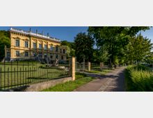 Gelbes historisches Gebäude mit reich verzierter Fassade und Statuen auf dem Dach, umgeben von einem gepflegten Garten mit einem schmiedeeisernen Zaun; daneben ein gepflasterter Geh- und Radweg, gesäumt von Bäumen und Grünflächen.