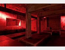 Ein industrieller Kellerraum aus Beton und Ziegelmauerwerk, beleuchtet von intensivem roten Licht. Der Raum enthält massive Pfeiler und Bänke und wirkt roh und unverkleidet, mit freiliegenden Leitungen und abgenutzten Oberflächen.