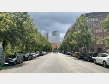 Baumgesäumte Straße mit parkenden Autos auf beiden Seiten; im Hintergrund ein modernes Hochhaus mit der Aufschrift „Park Inn“ und ein U-Bahn-Eingang. Auf beiden Seiten stehen mehrstöckige Wohn- und Geschäftshäuser.