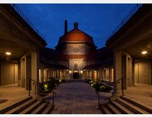 Ein historisches Gebäude mit einer achteckigen Kuppel im Zentrum, umgeben von symmetrischen Säulengängen. Der beleuchtete Innenhof mit Treppen, Pflanzen und gepflastertem Boden wird von warmem Licht erhellt, während der Hintergrund von der Abenddämmerung geprägt ist.