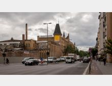 Straßenansicht mit einem historischen Backsteingebäude im Hintergrund, das durch Türme und Schornsteine hervorsticht. Im Vordergrund befinden sich eine belebte Fahrbahn, parkende Autos und Fußgänger, flankiert von modernen Wohnhäusern auf der rechten Seite.