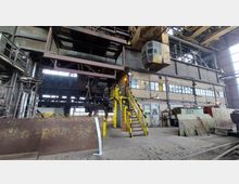 Innenansicht einer großen Industriehalle mit Stahlkonstruktionen und gelben Sicherheitsgeländern. Zu sehen sind hohe Wände aus Ziegeln, industrielle Maschinen und eine Hebevorrichtung mit einer Kabine.