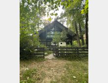 Ein modernes Haus aus dunklem Holz mit einem großen Giebeldach und einer Glasfront, umgeben von dichtem Grün und Bäumen. Vor dem Haus befindet sich ein niedriger Holzzaun mit einem offenen Tor und einem kurzen Weg, der zum Eingang führt.