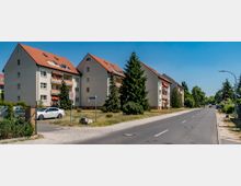 Mehrfamilienhäuser mit hellen Fassaden und roten Satteldächern entlang einer ruhigen Straße mit Gehwegen. Im Vordergrund sind parkende Autos, ein Schild mit einer Tempo-30-Zone und begrünte Vorgärten mit Bäumen sichtbar.