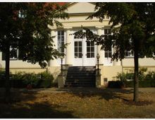 Historisches Gebäude mit symmetrischer Fassade, weißen Säulen und einer zentralen Treppe, die zu einer Glastür mit Sprossenfenstern führt. Umgeben von Bäumen und gepflegtem Gartenbereich.