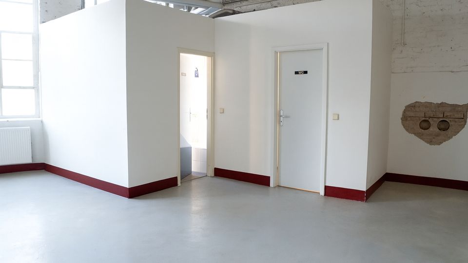 Innenraum einer industriellen Halle mit glattem grauem Boden, hohen Fenstern und sichtbaren Deckenrohren. In der Mitte steht ein weißer, boxförmiger Einbau mit zwei Türen, eine davon mit „WC“-Schild, sowie rotem Sockelabschluss.