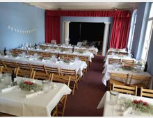 Ein festlich dekorierter Saal mit blau gestrichenen Wänden und roten Vorhängen, ausgestattet mit langen, weiß gedeckten Tischen und Holzstühlen. Auf den Tischen stehen Blumengestecke, Kerzenständer und Weinflaschen; im Hintergrund ist eine kleine Bühne mit roten Vorhängen sichtbar.