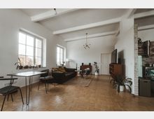 Ein geräumiges Wohnzimmer in moderner Einrichtung mit weiß gestrichenen Wänden, großen Fenstern, hellen Holzfußböden und sichtbaren Deckenbalken. Der Raum enthält ein Sofa, einen Esstisch mit Stühlen, Pflanzen, ein Sideboard, eine Stehlampe und eine offene Küche mit einer gemauerten Trennwand.