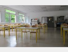 Ein einfacher Klassenraum mit rechteckigen Tischen und Stühlen aus Holz mit gelben Metallbeinen. Große Fenster auf der linken Seite lassen Tageslicht herein, und im Hintergrund befinden sich eine geschlossene Tür, Regale und eine aufgestellte Staffelei.