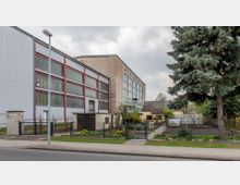 Industriegebäude mit großen Fensterflächen und einer angrenzenden kleinen Wohnsiedlung, umgeben von einem gepflegten Garten mit Blumenbeeten, einem Gewächshaus und einem großen Nadelbaum im Vordergrund. Ein Bürgersteig und eine Straße verlaufen vor dem Grundstück.