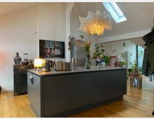 Moderne Küche mit grauer Kücheninsel, darauf eine Vase mit Blumen und Küchengeräte. Im Hintergrund helle Wände, ein Metall-Kühlschrank, Pflanzen, ein Kunstbild und eine große, weiße, skulpturale Lampe sowie Tageslicht durch ein Dachfenster.