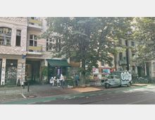 Straßenszene in einer urbanen Umgebung mit historischen Gebäuden, Cafés und Straßenkunst. Vor einem Café mit grünem Vordach ("Chestnut Coffee") stehen Tische und Stühle unter einem großen Baum; daneben ist ein graffitibedecktes Fahrzeug und mehrere Fahrräder geparkt.