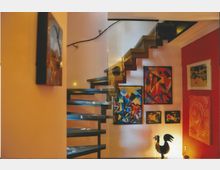 Moderner Innenraum mit einer Holztreppe und Glasgeländer, die in ein Obergeschoss führt. An den farbigen Wänden sind mehrere abstrakte Gemälde angebracht, und im Vordergrund steht eine dekorative schwarze Hahnskulptur.