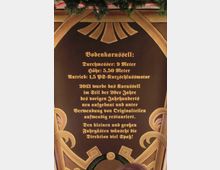 Holztafel mit goldener, verzierter Schrift im historischen Stil, die Informationen über ein Bodenkarussell gibt, einschließlich Durchmesser, Höhe und Antrieb. Dekorative Holzrahmen und Tannenzweige sind sichtbar am oberen Rand.