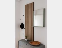 Minimalistisch eingerichteter Eingangsbereich mit einer grauen Kommode, darauf eine kleine Pflanze in einem Topf und ein Korb. An der Wand ein vertikales Holzpaneel, ein runder Spiegel mit Ablage und einige schwarze Wandhaken, von denen einer einen Hut trägt.