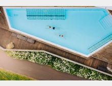 Rechteckiger Außenpool mit hellblauem Wasser, umgeben von einer Holzterrasse mit Liegen. Im Vordergrund ein Gehweg und blühende Hecken; im Pool schwimmen zwei Personen.