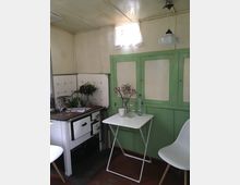 Rustikale Küche mit einer altmodischen, weißen Kochstelle und grünen Holzschranktüren. Ein kleiner weißer Klapptisch mit Dekorationen wie frischen Blumen, einer Kerze und einer Wasserflasche steht daneben, vor einer einfachen Einrichtung mit Pastelltönen.